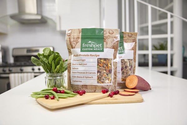 two bags of Freshpet Custom Meals on a white countertop surrounded by fresh ingredients: green beans, cranberries, sweet potato, and spinach