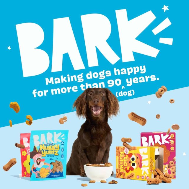 A dark brown dog sits in front of a bowl overspilling with dog treats, with packages of brightly colored treats all around. The image reads in big white letters: "BARK: Making dogs happy for more than 90 (dog) years."