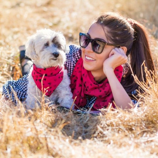 A pretty young woman with a brunette ponytail and sunglasses is gazing lovingly at a fluffy white dog as they both wear matching red bandanas