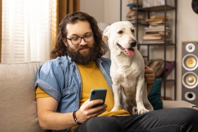 A man with glasses, long hair and a scruffy beard sits on a couch with a white mixed breed dog on his lap while looking at his cell phone