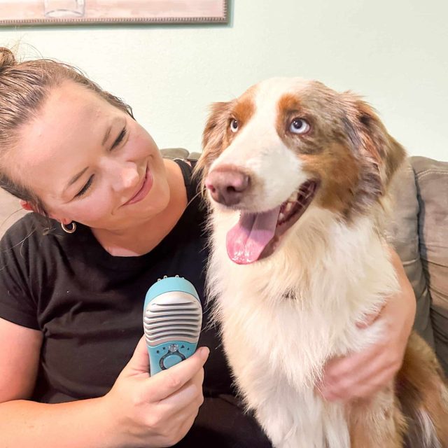 A smiling woman sits with her arm around an Australian Shepherd dog, and holds a MedcoVet Luma device in her right hand.