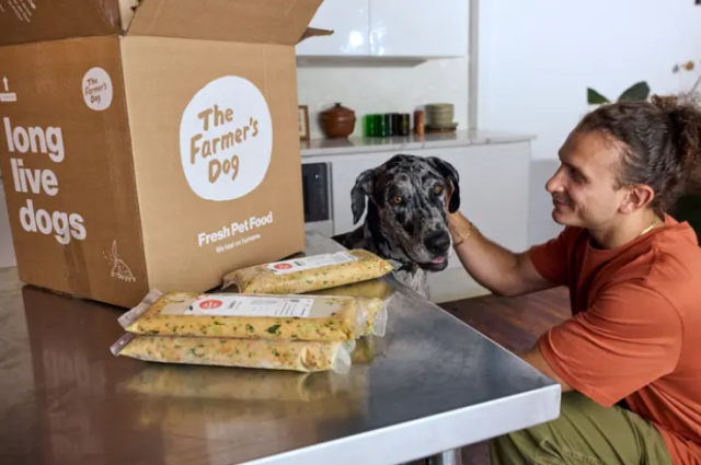 A Farmer's Dog delivery box and several packets of fresh dog food sit on a table as a man pets his Great Dane.