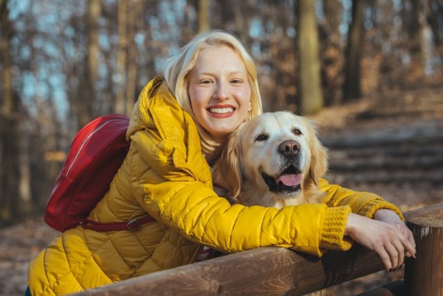 A blond woman in a yellow puffer jacket stands with her arms around a senior Golden Retriever.